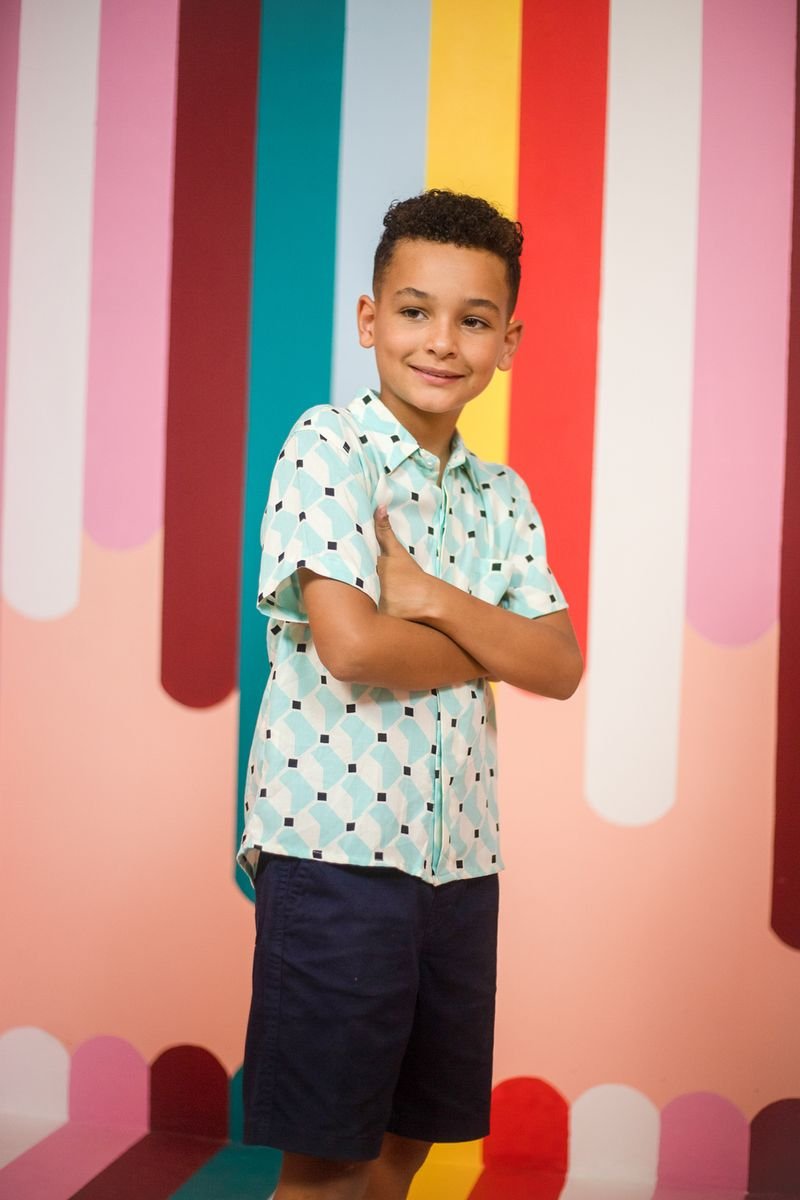 Child in a light blue patterned short-sleeve shirt and navy shorts in front of a colourful striped wall